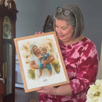A silver-haired woman in a pink tie-dye top holds a light oak-framed watercolor-style portrait of a woman and a toddler