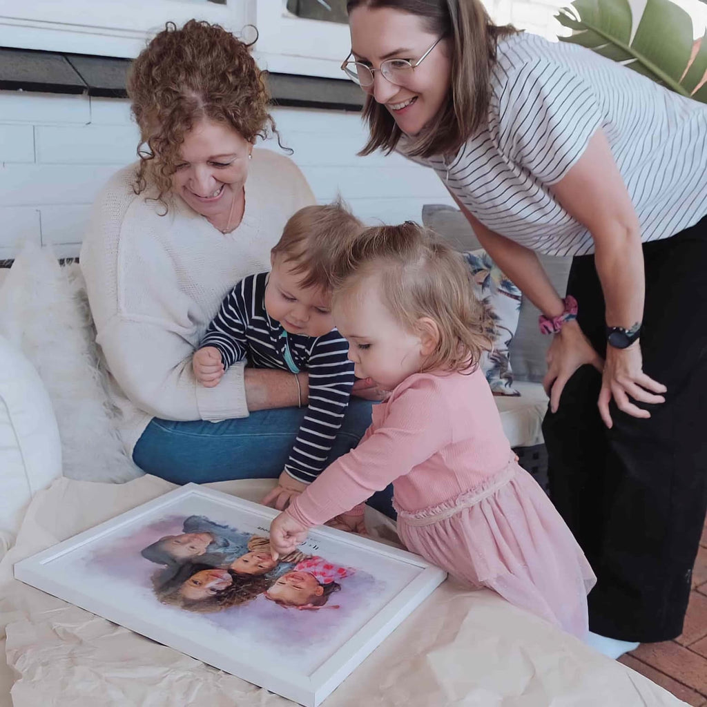 Two women and two toddlers look at a white-framed family portrait unwrapped on a table