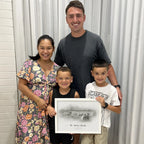 A woman, a man and two boys smile for the camera; a boy holds a white-framed sketch-style portrait of a house with a family