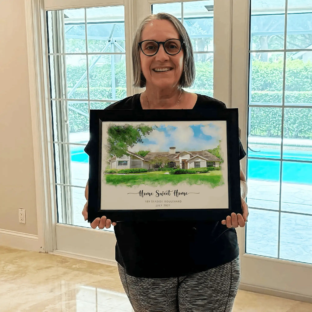 A smiling woman holds a framed watercolor-style portrait of a single-story house titled 'Home Sweet Home'