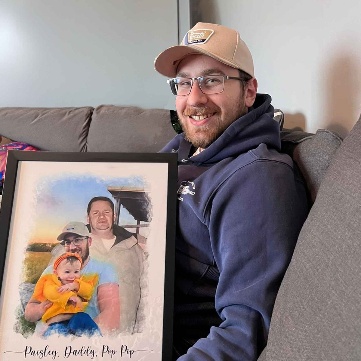 A smiling man in a cap holds a black-framed watercolor-style portrait of a man and a toddler in a yellow outfit