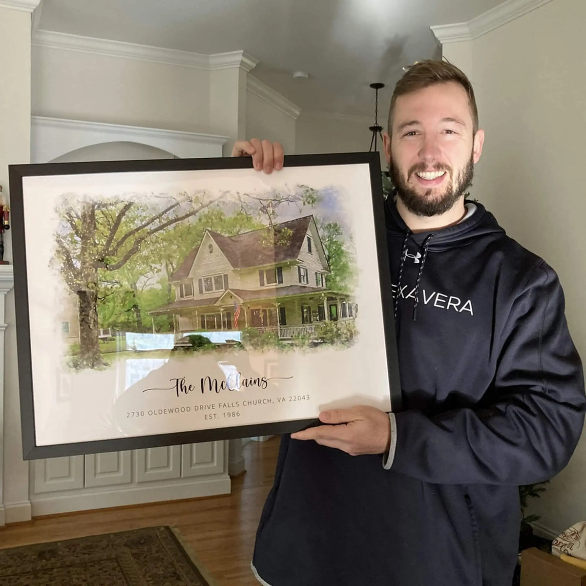 A smiling man holds a black-framed watercolor-style house portrait of a pale yellow house, titled 'The McClains'